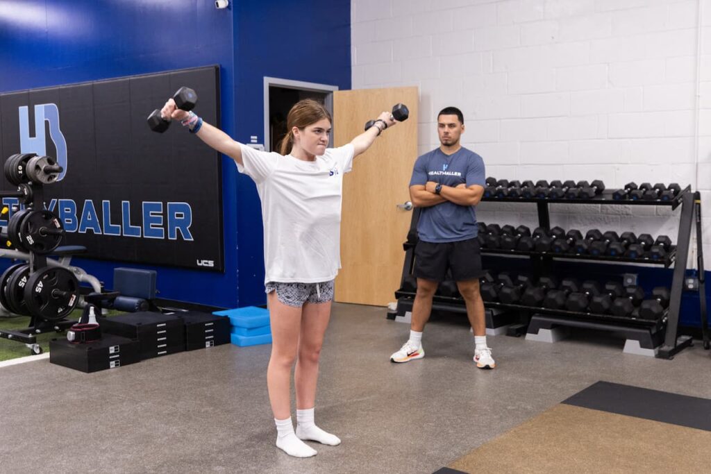 Woman lifting dumbbells in gym with trainer watching.