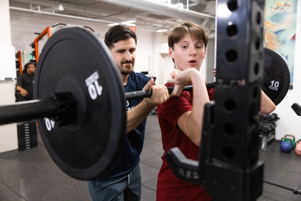 Trainer assists youth during barbell lifting exercise.