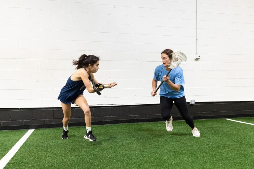 Two women playing lacrosse indoors.