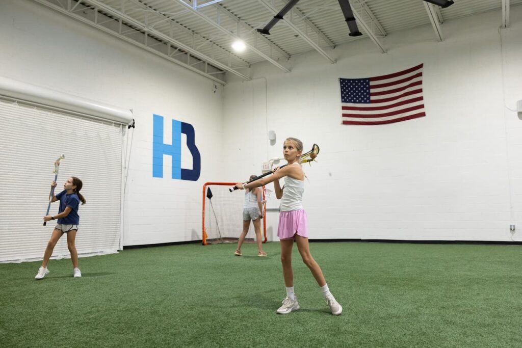 Children practicing lacrosse indoors on turf field.