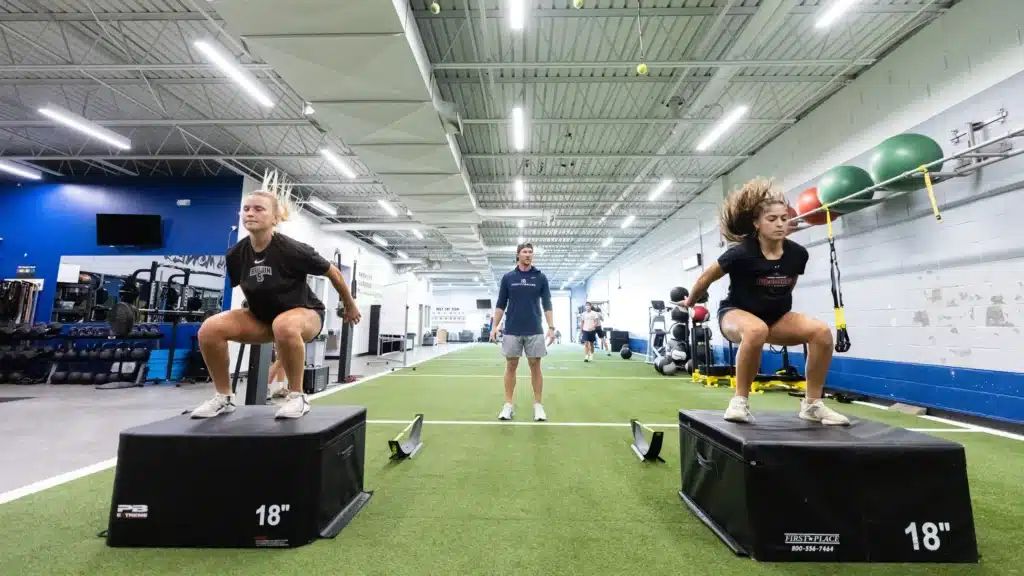 Two women performing box jumps in gym.