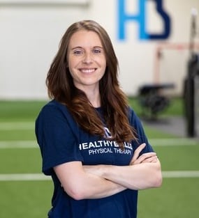 Smiling woman in gym setting, physical therapy shirt.