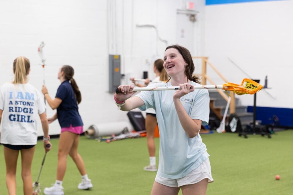 Girls practicing lacrosse indoors on green turf.