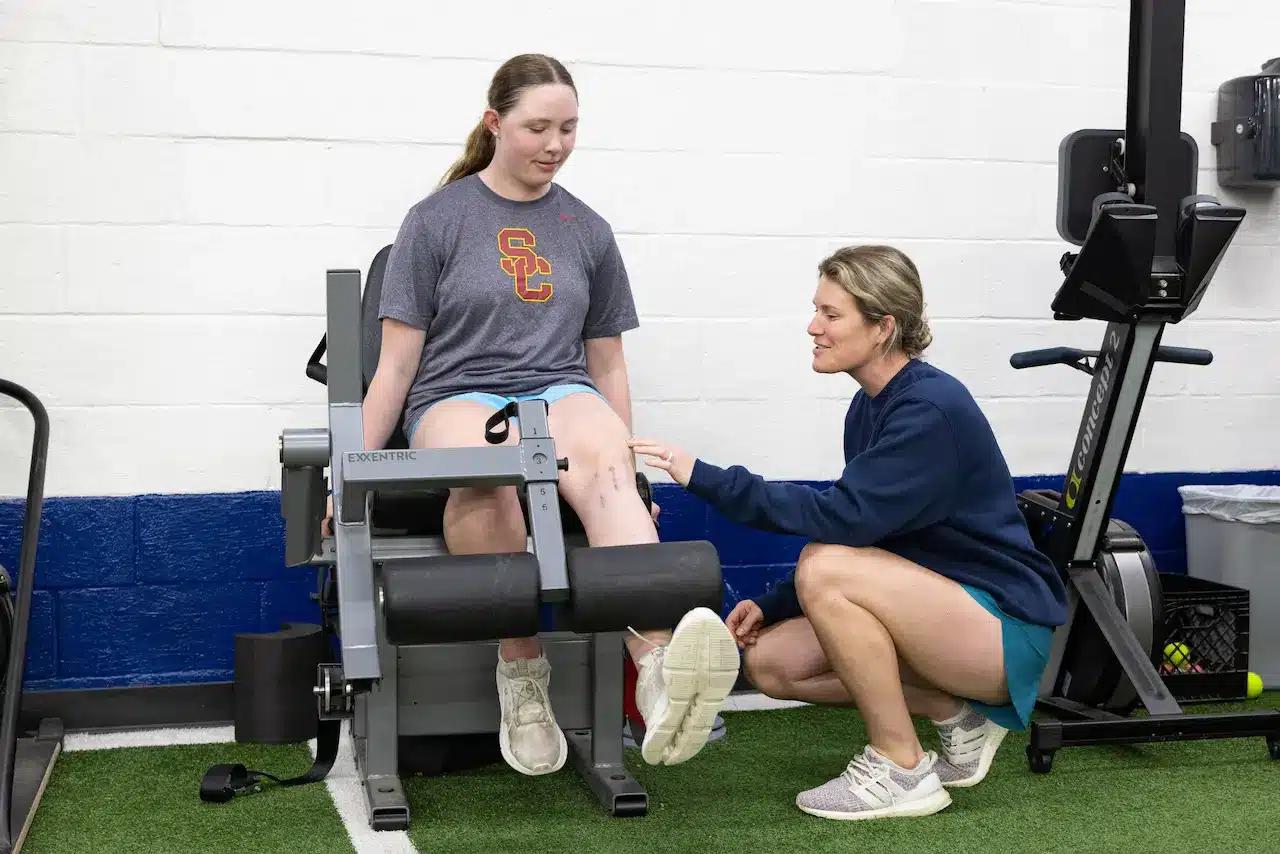 Trainer assists woman on leg exercise machine.