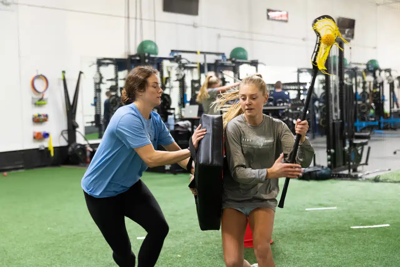 Women training indoors with lacrosse stick and pad.
