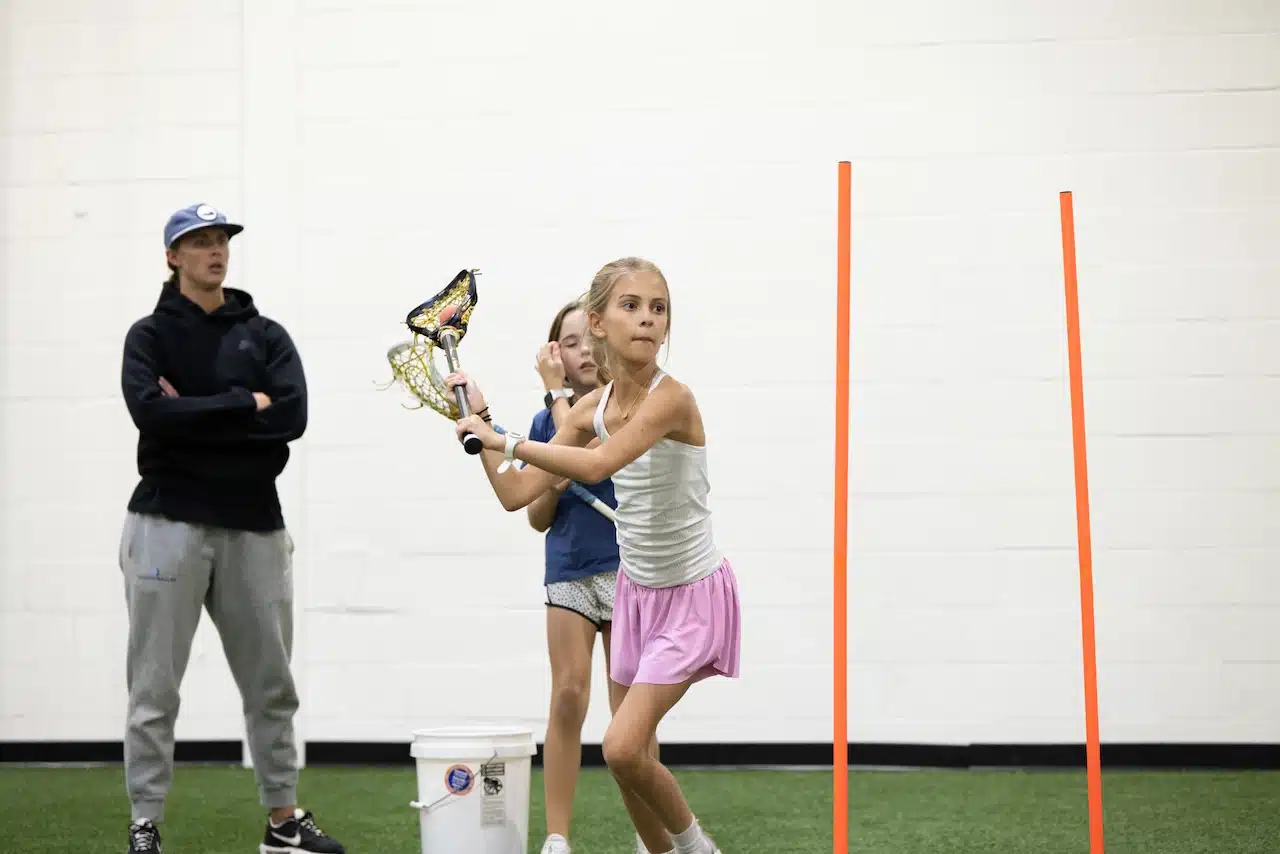 Girls practicing lacrosse indoors with coach overseeing.